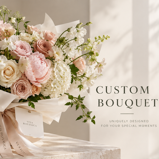 A beautifully arranged custom bouquet of pink and white flowers with green foliage, wrapped in elegant cream paper and a matching ribbon, displayed on a marble surface with soft window shadows. Text on the right reads 'CUSTOM BOUQUET'.