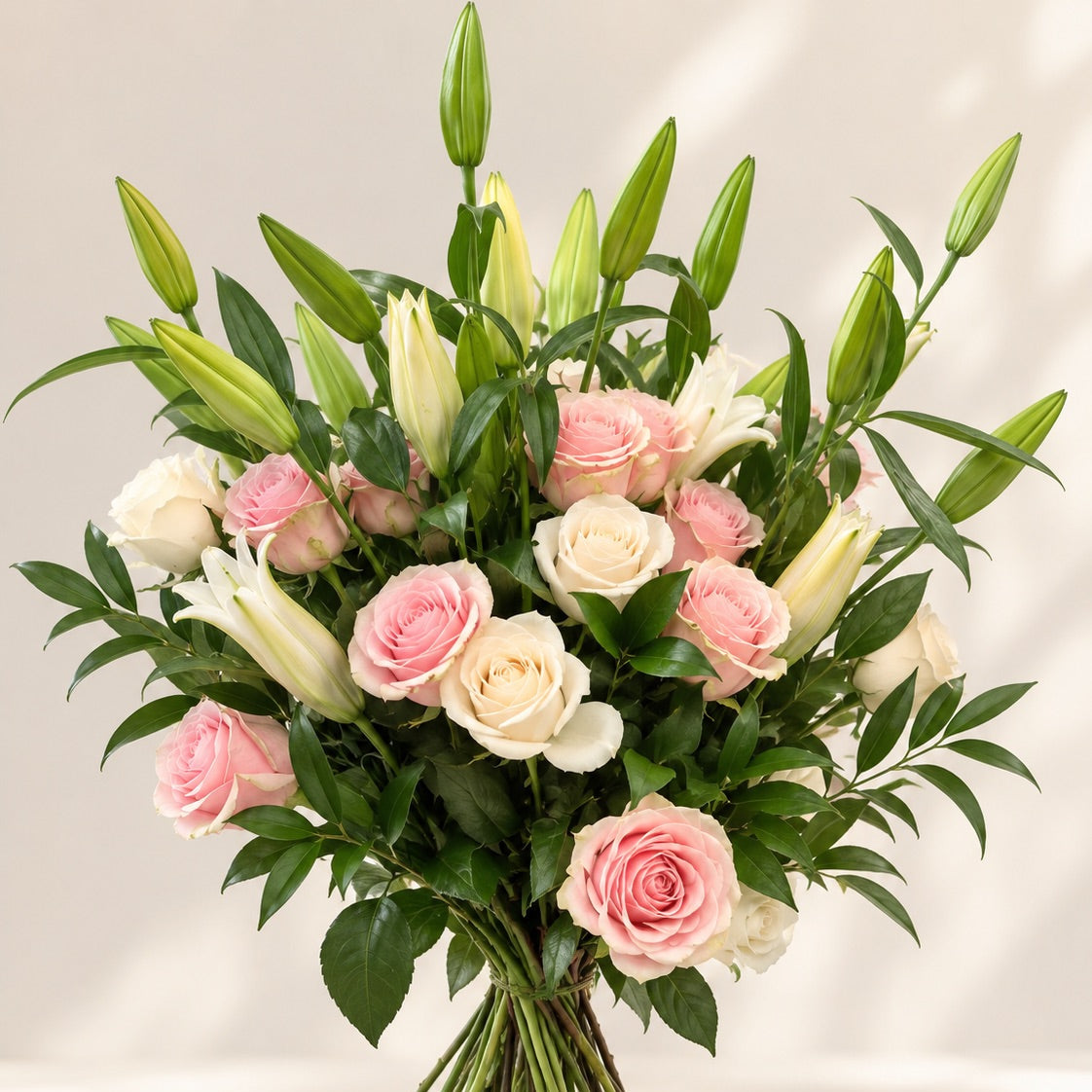 A vibrant floral bouquet featuring pink and cream roses, white lilies, and green lily buds with lush green leaves, arranged in a dense bundle and set against a soft beige background with subtle shadows.