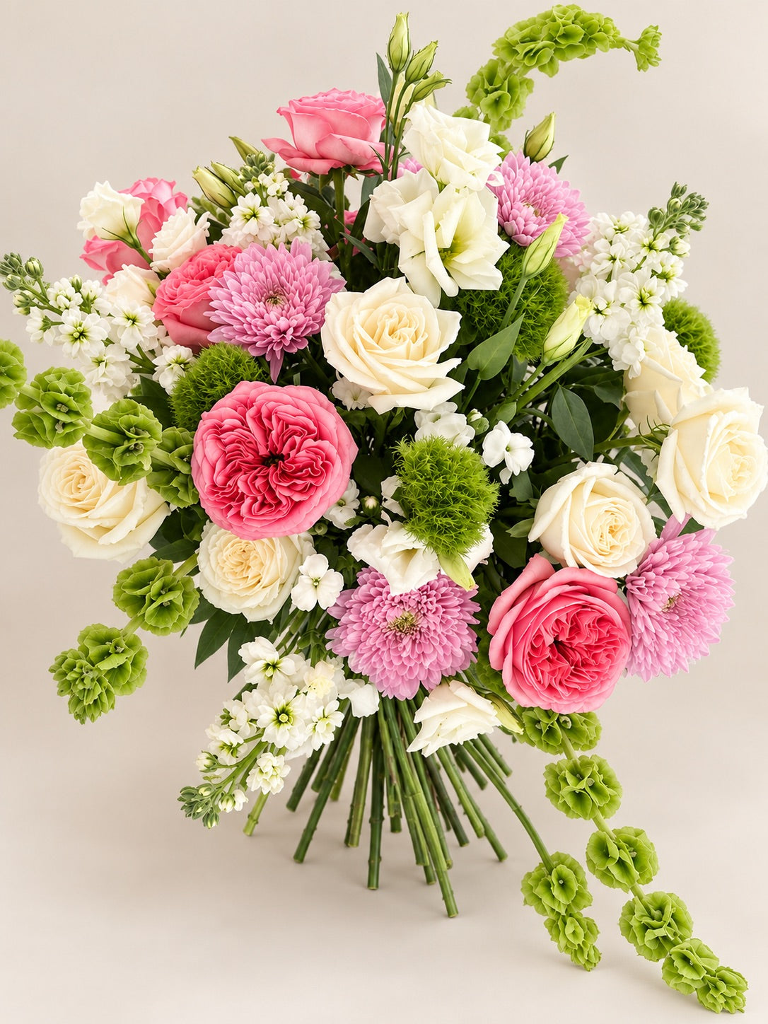 A vibrant floral bouquet featuring pink and cream roses, chrysanthemums, green hydrangeas, and white filler flowers, arranged against a solid light beige background.